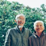 A happy elderly couple smiles warmly outdoors against a vibrant green background.