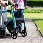 Senior adults walking with a rollator along a park path on a sunny day.