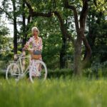 Elderly woman in floral dress standing with a bicycle in a vibrant, green park.