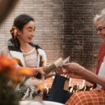 Two women joyfully exchanging gifts by a warm indoor fireplace.